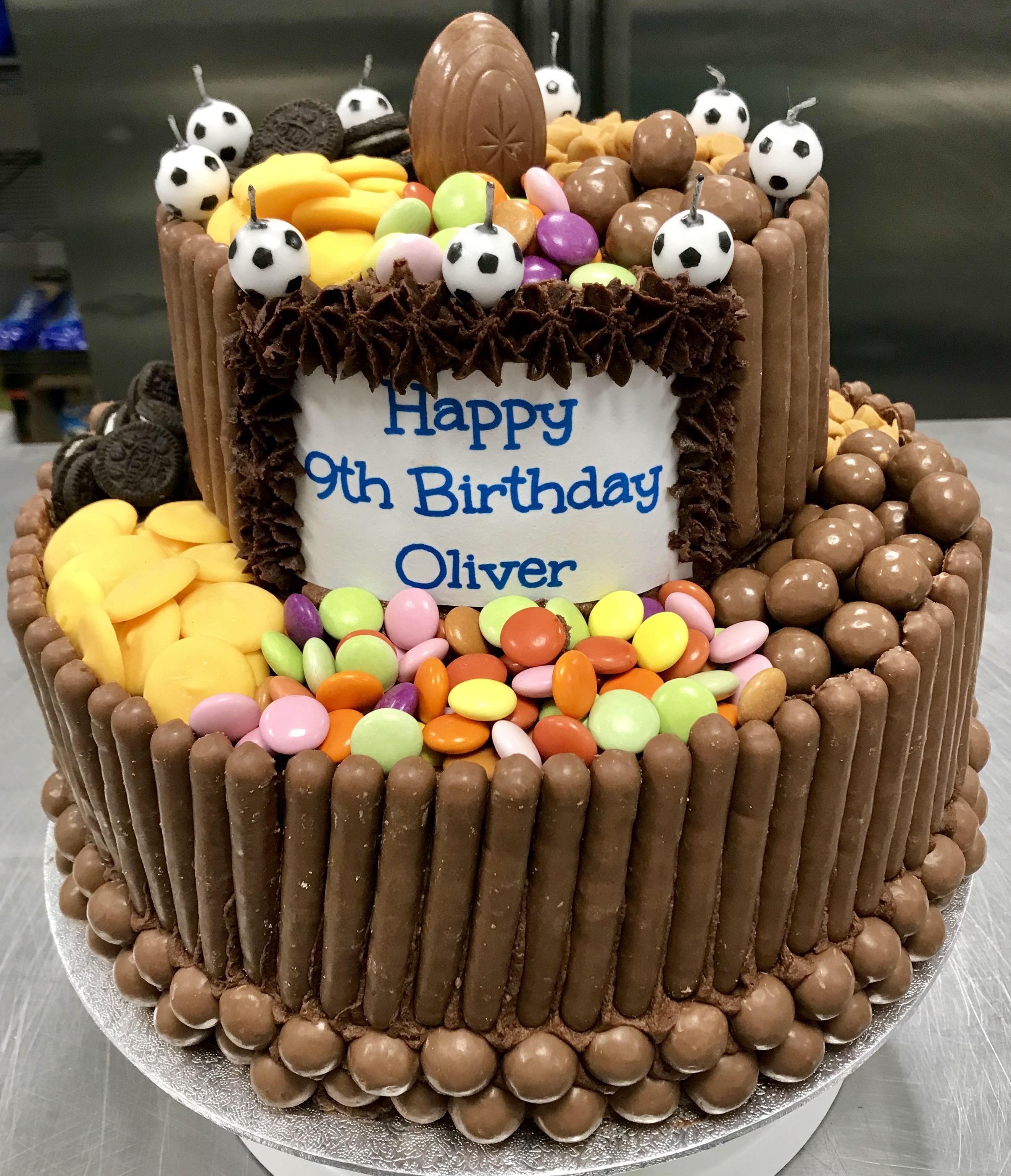 A two-tiered Chocolate Blackout Cake adorned with various sweets and chocolates. The sides are lined with chocolate fingers and Maltesers, while the top is decorated with colorful chocolate candies, chocolate soccer balls, and a large chocolate egg. A plaque reads, 'Happy 9th Birthday Oliver.'